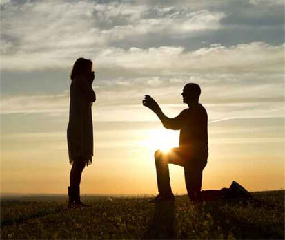 A man is kneeling down to propose to a woman at sunset.