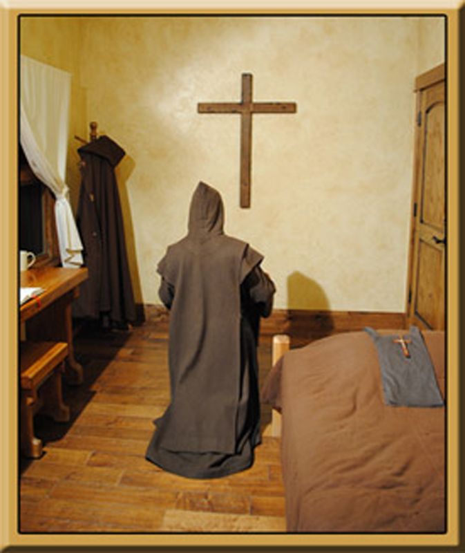 A monk in a room with a bed and a cross.